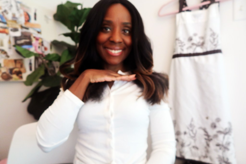 A smiling person dressed in a white shirt, playfully gesturing with their hand while seated in a bright room with plants and a hanging garment in the background.