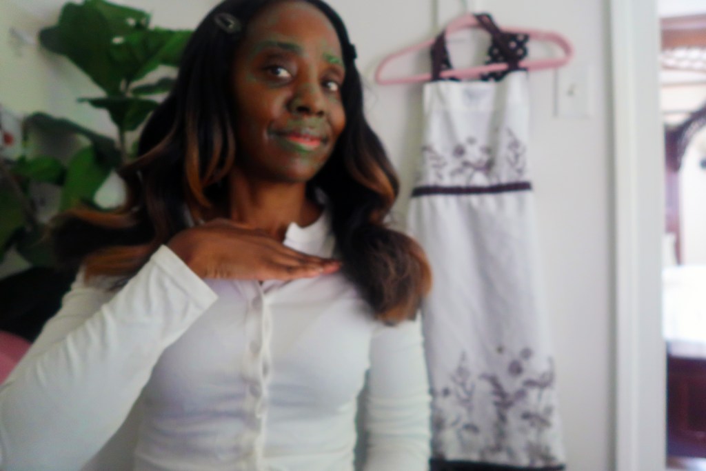 A person displaying a homemade face mask with green clay on their face, smiling while gesturing with their hand, indoors with a plant and a dress hanging in the background.