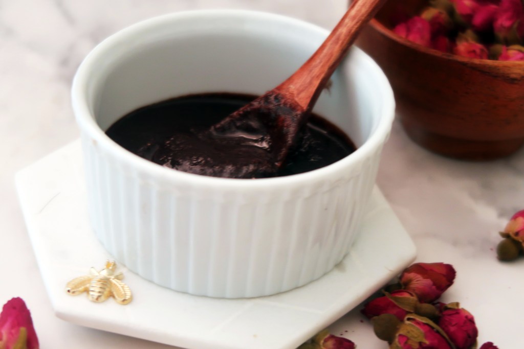 A small white bowl filled with a dark brown scrub, with a wooden spoon resting inside, surrounded by dried rose petals and a wooden bowl.