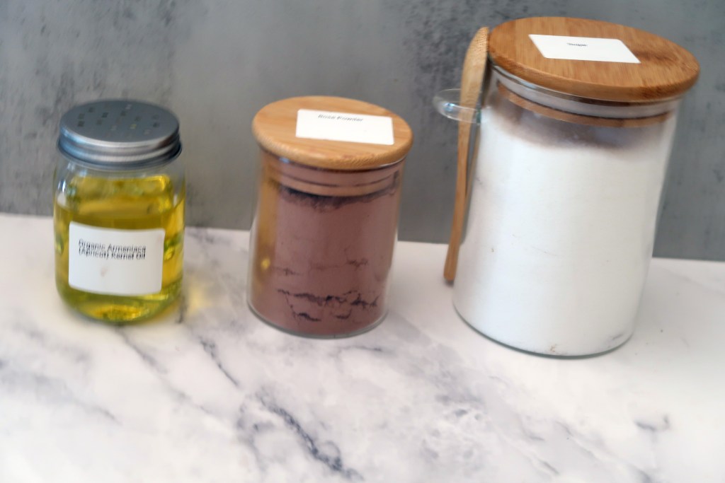 Three jars with labels: one contains apricot oil, another has rose powder, and the third is filled with granulated white sugar.