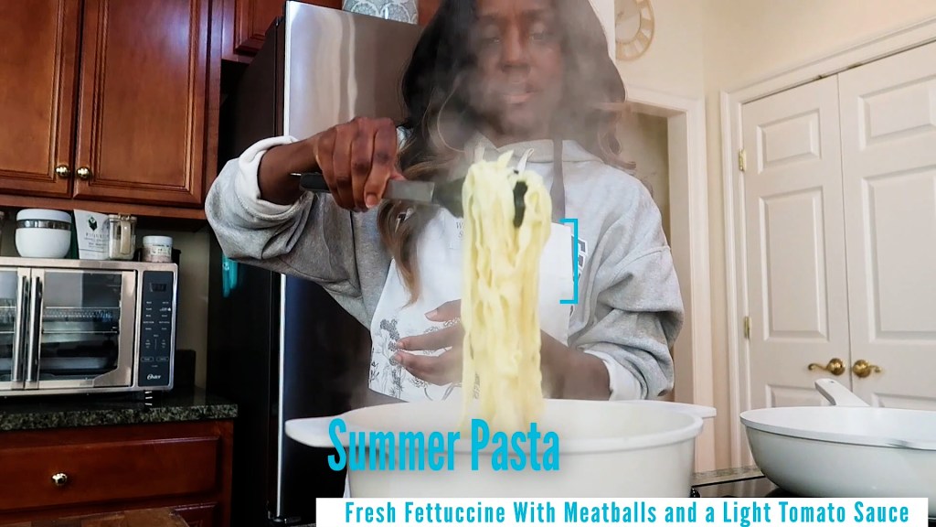 A person stirring fresh fettuccine pasta in a bowl, with steam rising in a kitchen setting.