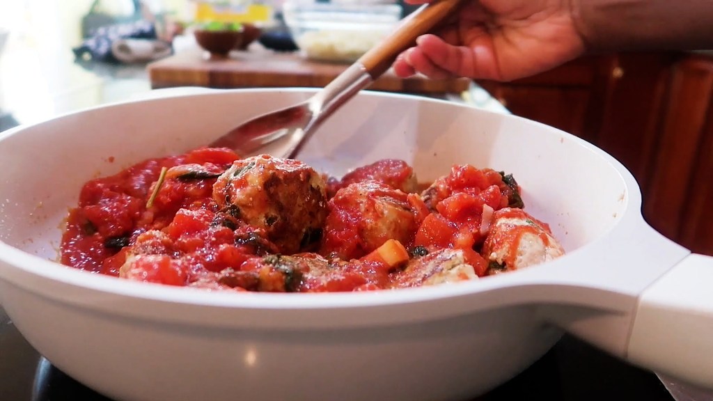 A person stirring turkey meatballs in a white bowl with tomato sauce, garnished with herbs, in a kitchen setting.