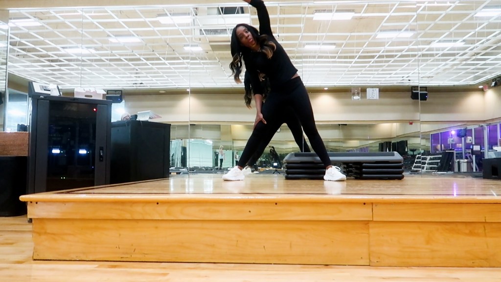 A person performing a stretching exercise on a wooden stage in a gym, surrounded by mirrors and fitness equipment.