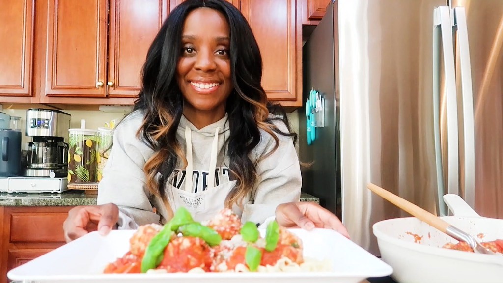 A person smiling while holding a plate of spaghetti topped with meatballs and garnished with basil in a kitchen setting.