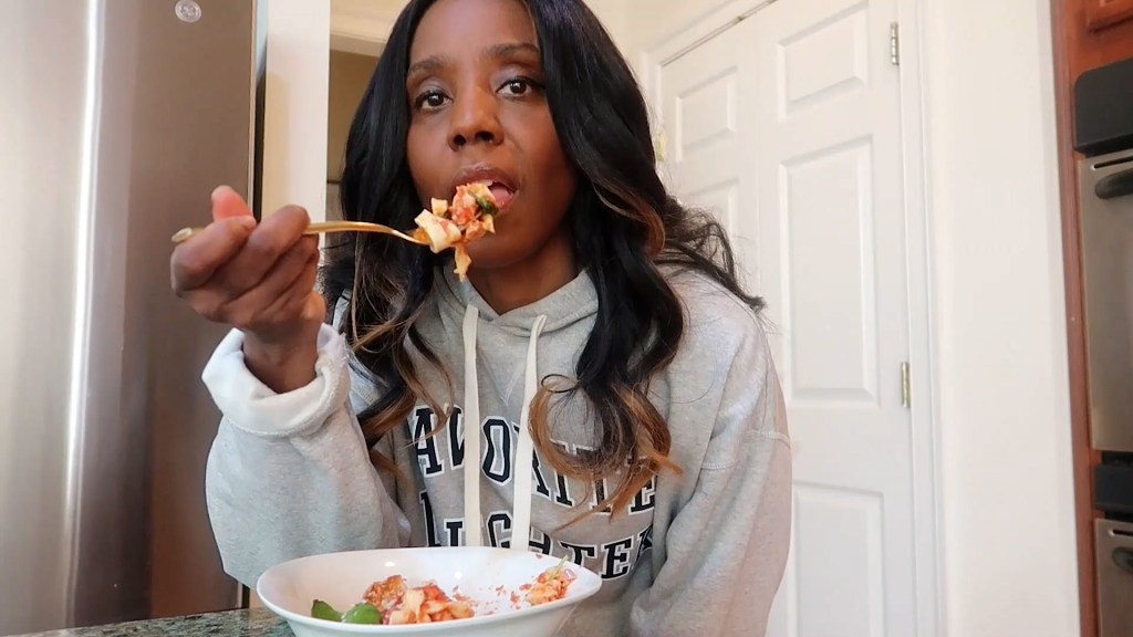 A person sitting at a kitchen table eating pasta with a fork, wearing a gray hoodie.